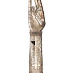 43. Reliquary-Gift of the Estate of Aghavny Demirjian, 18th-19th century Armenian Museum of America, Boston, MA. The hand is an austere design of heavy brass without ornamentation, with the fingers joined in blessing. The thumb is broken and has been repaired. The forearm is a lighter brass material, and elaborately decorated. The inside of the arm displays an intricate hammered floral design surrounding a simple cross. The outside of the forearm has three separate panels: a dove of the Holy Spirit with 2 angels, the sun and the moon (top), a cathedral with domed crosses (middle) and a haloed figure kneeling in a garden between two figures (Christ in the Garden of Gethsemane?). The top and bottom of the forearm are wrapped with twisted wires and mountings for encrusted gemstones, now lost. The base opens to permit access to the hollow center.